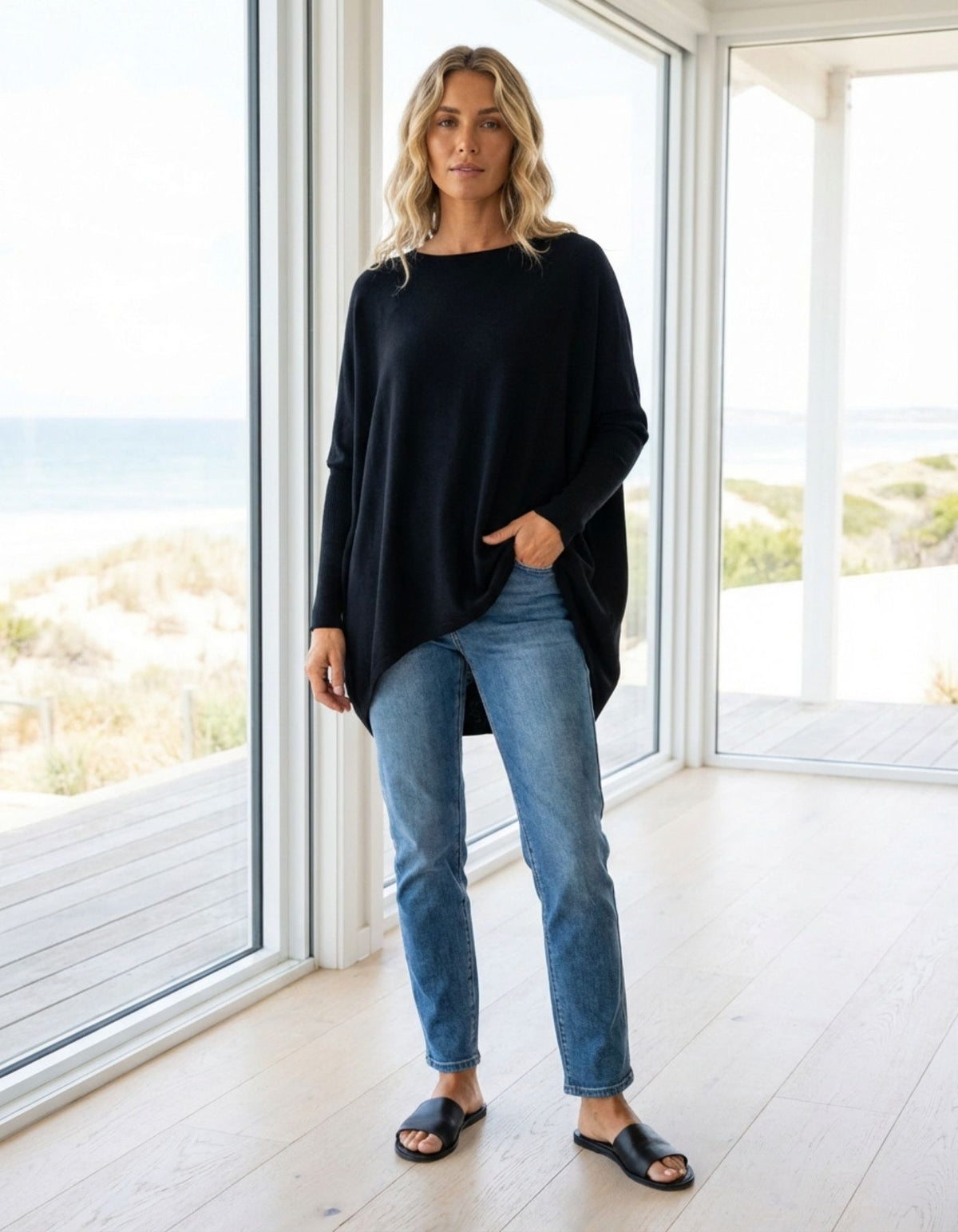 Woman wearing a black sweater and blue jeans standing in a bright room with large windows.