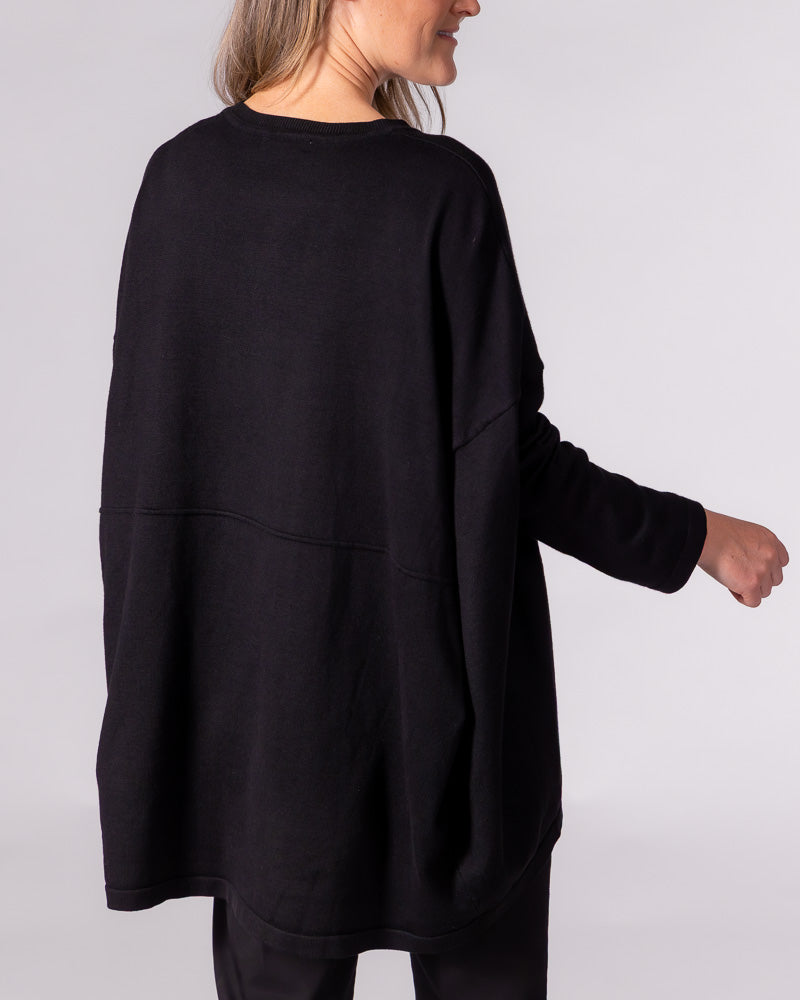 The back of a black oversized knit jumper displays a distinct horizontal seam, draped volume, and long fitted sleeves.