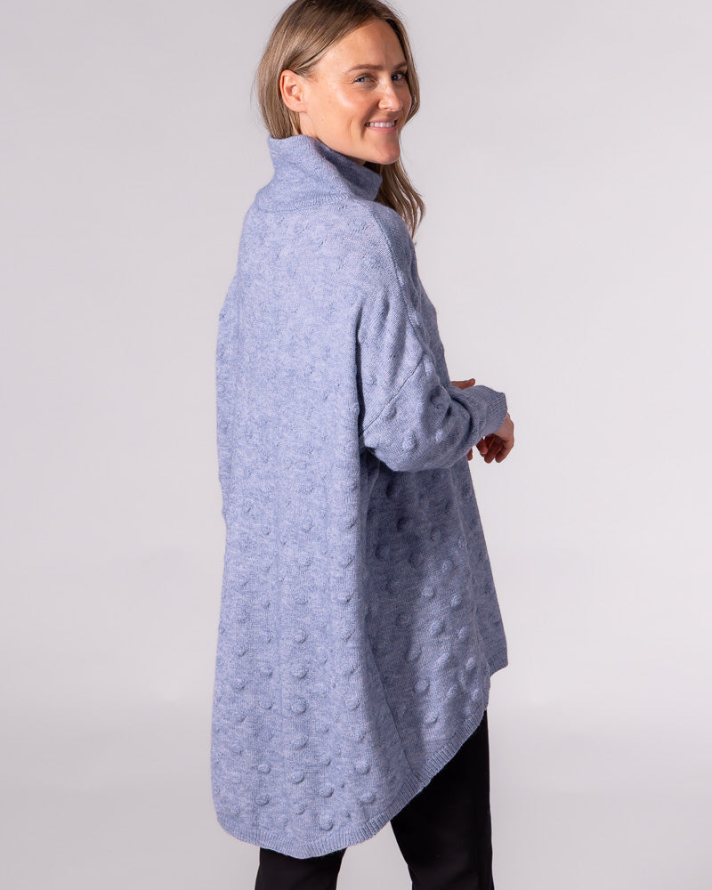Back view of a light blue oversized bubble knit jumper with a funnel neck, drop shoulders, and a scooped high-low hem.