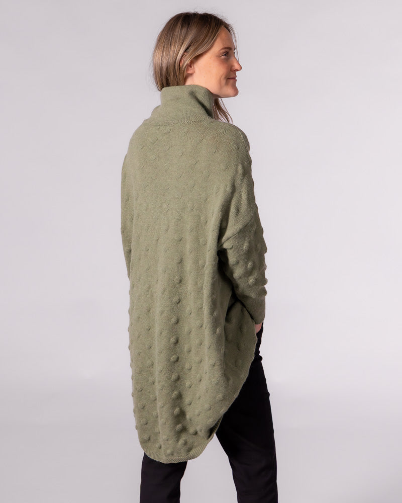 Back view of an oversized olive green jumper with a textured bubble knit, funnel neck, and long scooped back hem.
