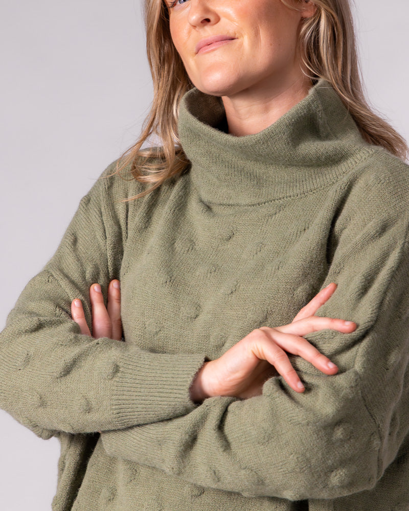 Olive green knit jumper featuring a ribbed funnel neck, relaxed drop shoulders and a raised bubble texture.