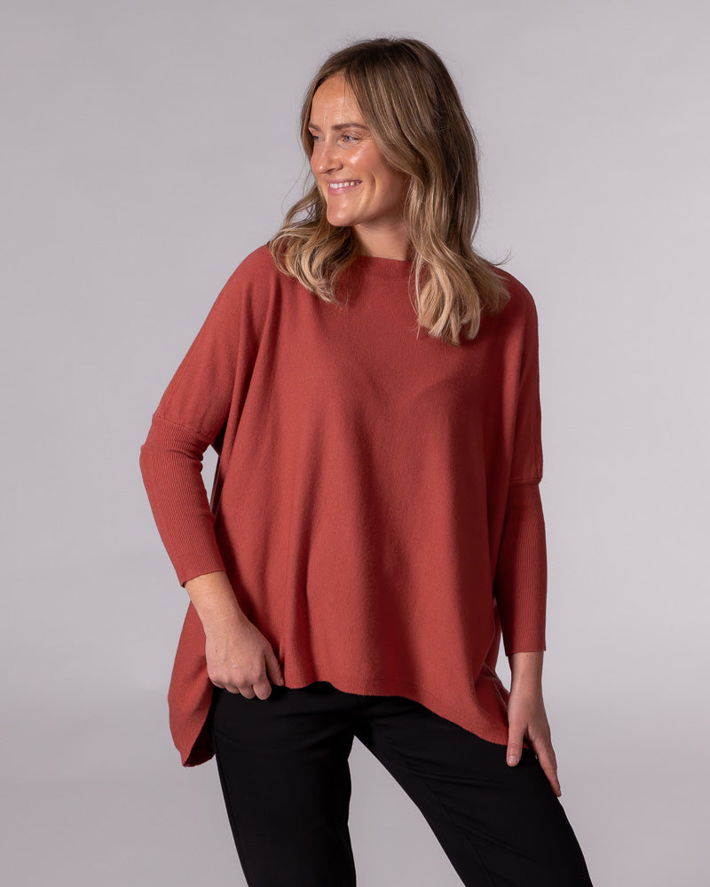 Oversized burnt orange cotton knit jumper featuring drop shoulders, fitted ribbed sleeves, and a draped hi-low hem.