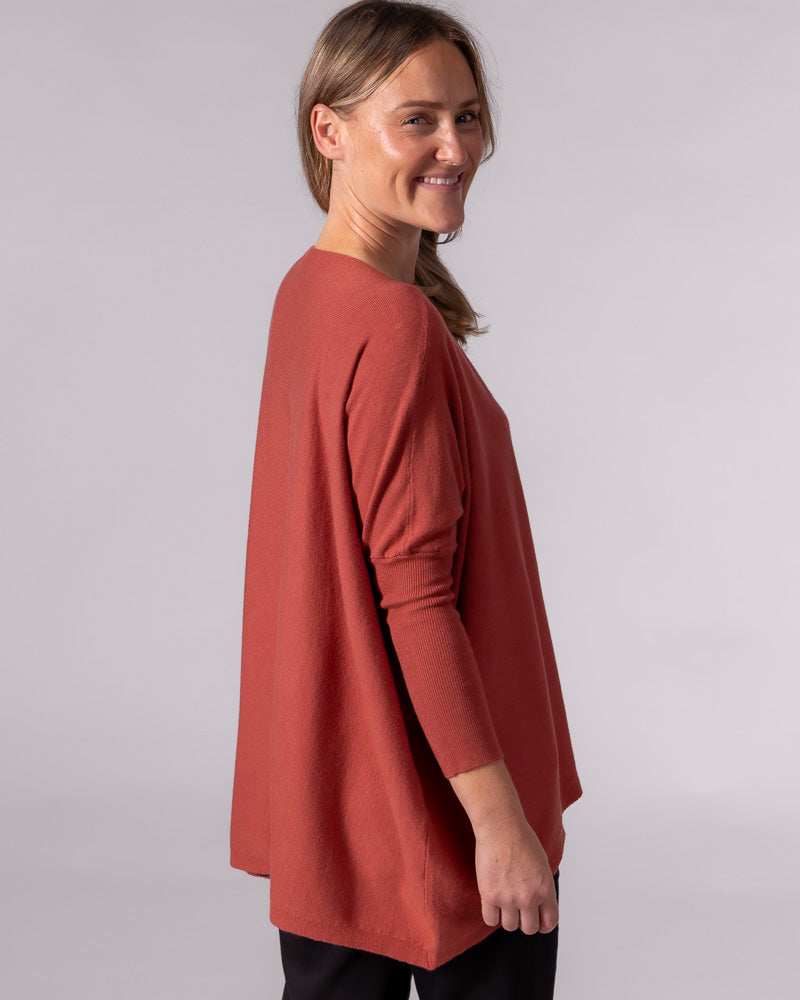 Burnt orange oversized cotton knit jumper showing a draped back, hi-low hem, drop shoulder, and fitted lower sleeve.
