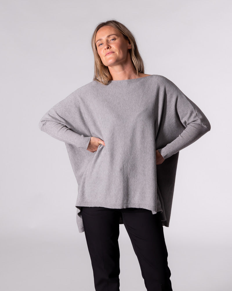 Front view of the grey cotton knitwear with hands on hips, showing the wide boat neckline and oversized drape.