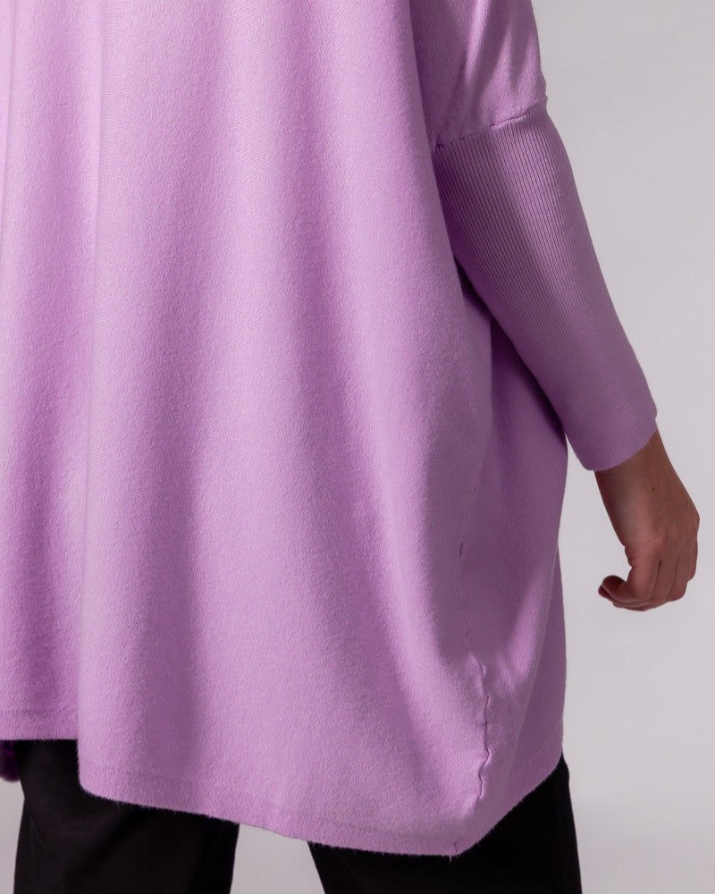 Oversized lilac cotton knit jumper displaying a draped silhouette, hi-low hem, and a fitted ribbed sleeve.
