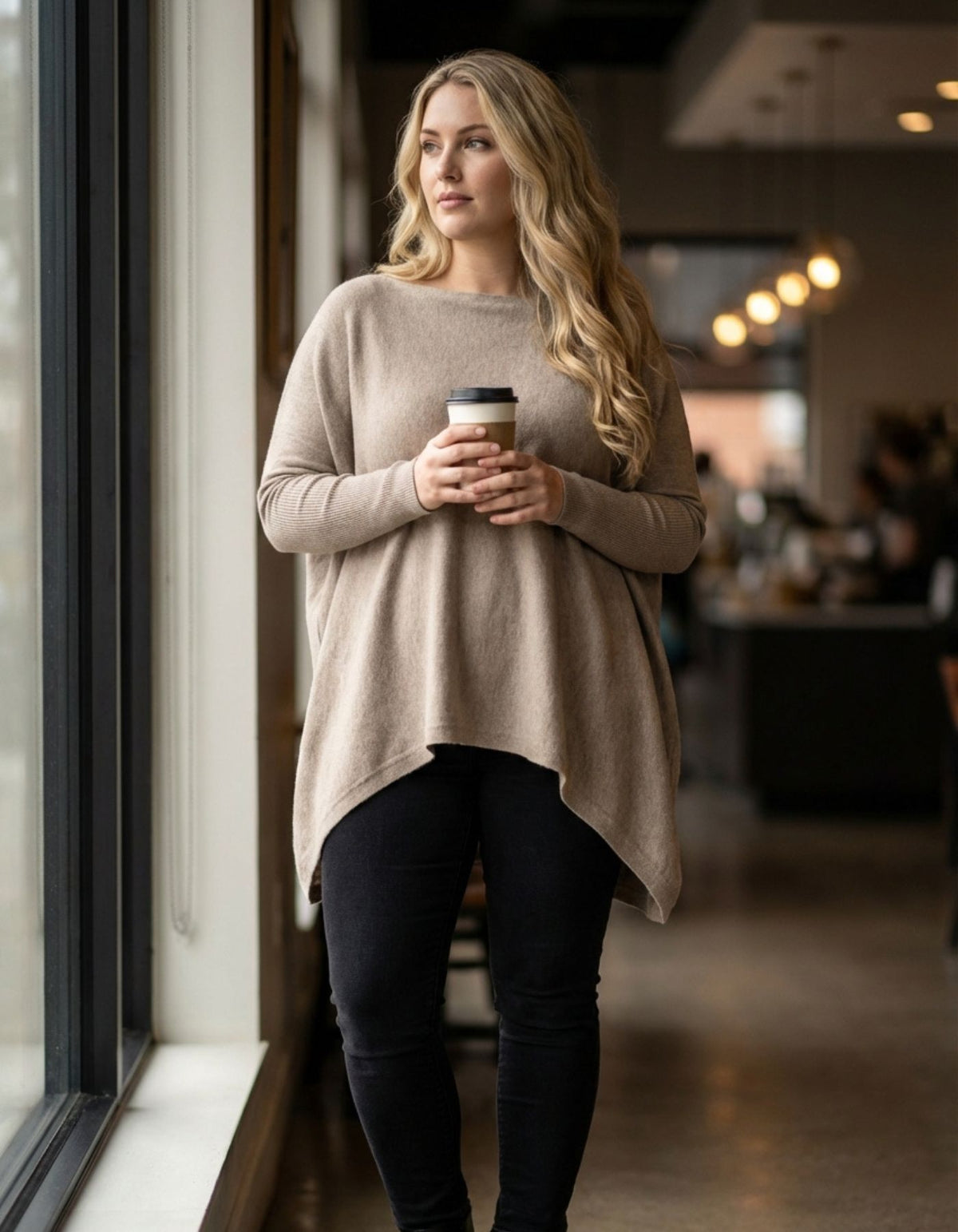 Full-length styling of the oversized Karma Kameleon taupe jumper paired with black denim jeans in a cafe setting.