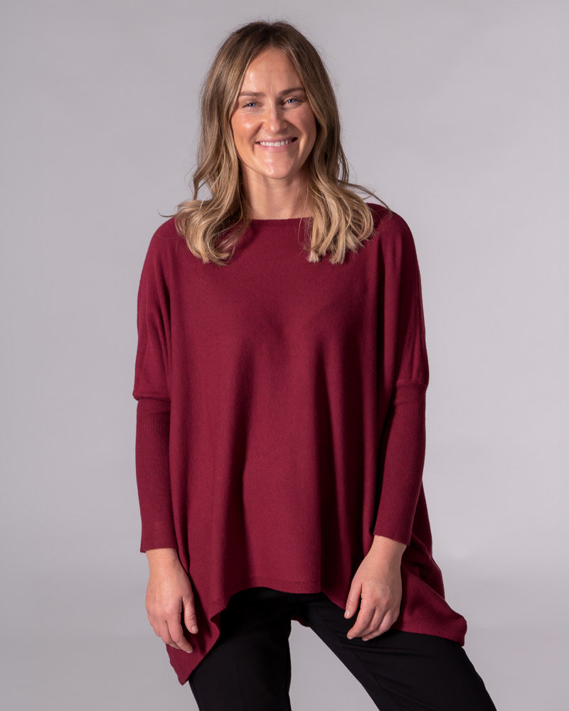 Oversized wine red cotton jumper featuring a wide boat neckline, drop shoulders, fitted sleeves, and a draped hi-low hem.
